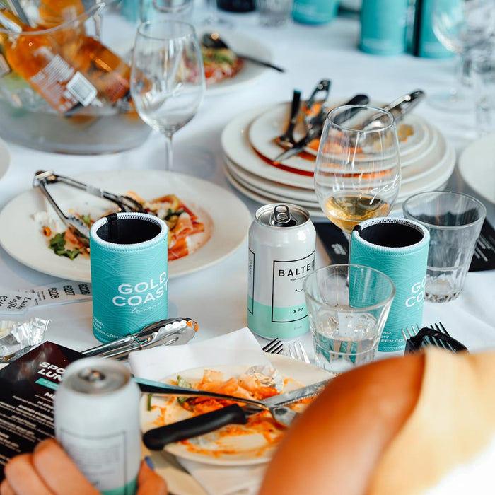 Custom Corperate Stubby Coolers holding aluminum cans on a white table covered with plates & glasses.