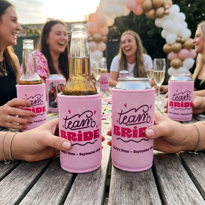 Custom Hens Stubby Coolers with 'Team Bride' text are held up on a wooden outdoor table, with people and champagne glasses in the background.